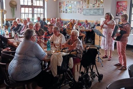 Eine Frau mit Flöte und eine Frau mit Akkordeon spielen in einem Saal, indem viele Menschen an Tischen sitzen.