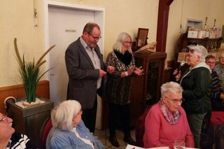 Im Vordergrund sitzen drei Menschen an einem Tisch. Im Bildhintergrund stehen der Ortsvorsitzende und seine Stellvertreterin und eine Frau, die sich mit einer Ansprache über Mikrofon bei ihnen bedankt.