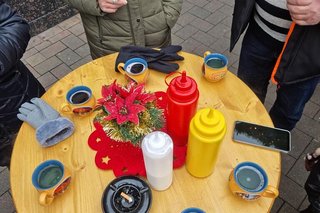 Ein Holzstehtisch von oben fotografiert. Darauf steht mittig ein kleiner Christstern und drumherum Glühweintassen und Tuben mit Senf und Ketchup.