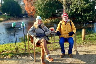 Gut gelaunt sitzen eine Frau und ein Mann mit warmen Jacken und Mützen in der winterlichen Abendsonne unter einem Baum am Ufer des zugefrorenen Schlossgrabens.