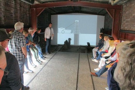 Menschen stehen und sitzen rechts und links in einem Innenraum des Zechengebäudes. Am Ende des Raums wird auf einer Leinwand ein Schwarzweiß-Film mit Aufnahmen aus der Betriebszeit der Zeche gezeigt.