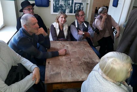Eine Gruppe Menschen sitzt gemeinsam um einen alten Holztisch herum und hört jemandem, der nicht im Bild zu sehen ist, zu.