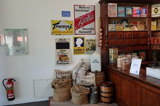 Alte Werbeschilder aus Blech hängen an einer Wand, darunter stehen Säcke aus Jute und Holzfässer. Rechts davon sieht man einen Teil des alten Apothekerregals mit Blechdosen und Produkten aus der damaligen Zeit