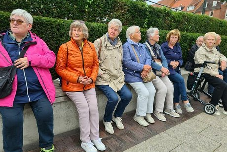 Eine Gruppe Menschen sitzt nebeneinander auf einer langen Bank vor einer Kirschlorbeerhecke.
