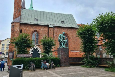 Kirchenvorplatz mit einer Bronzestatue des deutschen Lyrikers Emanuel Geibel auf einem Steinsockel.
