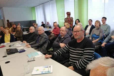 Menschen sitzen an langen Tischreihen und folgen einer Diskussionsrunde.