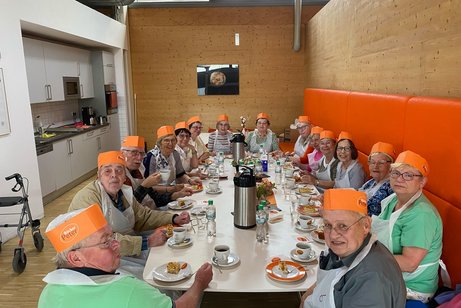 16 Personen sitzen an einem langen Tisch mit Sch&uuml;rzen und orangen B&auml;ckerhauben bekleidet.