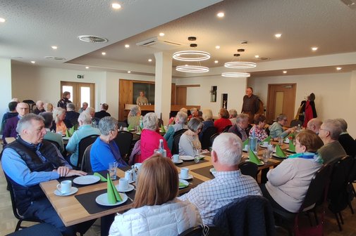Viele Menschen sitzen an gedeckten Tischen in einem Raum. An der rechten Seite steht ein Mann und spricht zu Publikum.