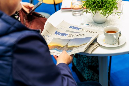 Ein Mensch in dukelblauem Anzug sitzt mit einer Tasse Kaffee an einem Tisch und liest eine Ausgabe der VdK-Zeitung