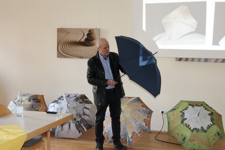 Ein Mann steht mit einem aufgespannten Schirm in der Hand vor anderen Schirmen und h&auml;lt einen Vortrag.