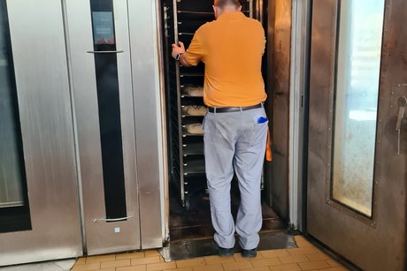 Besuch bei Bäckerei Peter. Ein Bäcker schiebt einen Wagen mit Backwaren in einen großen Backofen.