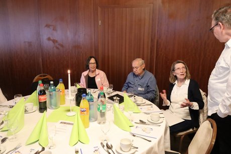 Drei Personen sitzen an einem runden Tisch und unterhalten sich mit einem stehenden Mann.