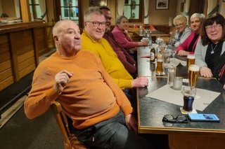 Eine Gruppe Männer und Frauen sitzen gemeinsam bei Getränken an einem langen Tisch.