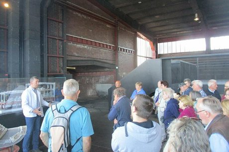 Eine Gruppe Menschen steht in der Halle der ehemaligen Kohlewäscherei und hört einem Mann zu, der etwas erzählt.