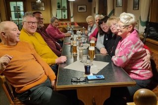 Eine Gruppe Menschen sitzt gemütlich in einer Gaststube an einem langen Tisch und redet und lacht.