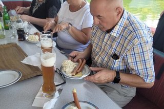 Zwei Frauen und ein Mann sitzen an Deck eines Ausflugsschiffs und genießen dort Bier, Kartoffelsalat und Würstchen.