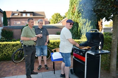 Ein Mann steht am Grill, zwei Männer mit Bierflaschen in der Hand daneben