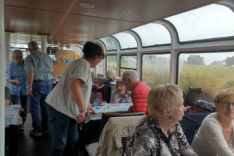 Menschen sitzen an Tischen auf dem Innendeck eines Schiffs. Durch große Panoramafenster sieht man das Kanalufer und grauen Himmel.