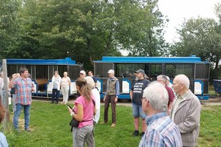 Die Besuchergruppe steht auf einer Wiese und lauscht den Erklärungen eines Mannes, der vor einem der Straußengehege steht. Im Hintergrund wartet die blauweiße Bimmelbahn.