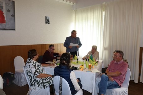 Jahreshauptversammlung 2026 Mehrere Personen sitzen an einem Tisch. Ein Mann steht und liest etwas von einem Blatt ab.