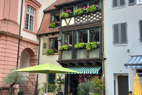 In der Bildmitte ein altes mit Blumen geschmücktes Fachwerkhaus; rechts und links sind neuere Gebäude zu sehen, die das Fachwerkhaus einrahmen.