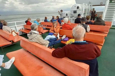Menschen sitzen auf orangen Plastikbänken auf dem Außendeck einer Fähre.