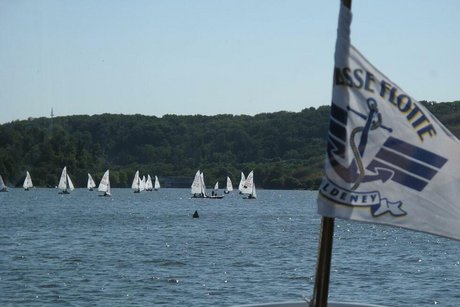 Zahlreiche Segelschiffe kreuzen über den Baldeneysee. Im Hintergrund die bewaldete Ruhranhöhe, im Vordergrund flattert eine Fahne der Weißen Flotte im Wind.