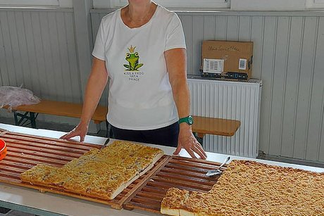 Eine Frau steht hinter einem Tisch, auf dem zwei riesige Bleche Streuselkuchen stehen, und lächelt in die Kammera.