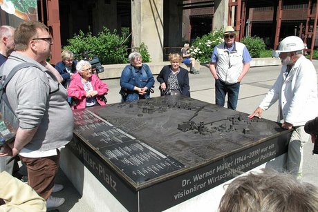Die Reisegruppe steht um einen barrierefreien Geländeplan aus Bronzeguss herum. Rechts im Bild steht der Museumsführer mit weißem Helm und weißer Bergmannsjacke.
