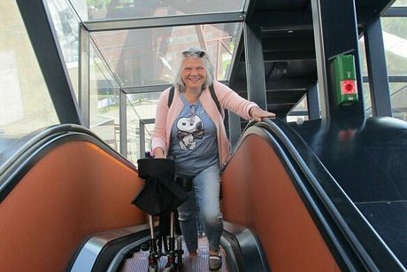 Blick von oben auf die mit Glas verkleidete Rolltreppe, auf der gerade eine Frau mit zusammengeklapptem Rollator nach oben fährt.
