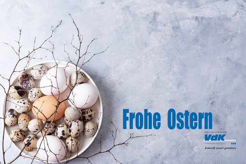 Eine Schale mit naturfarbenen kleinen und gro&szlig;en Eiern und Zweigen auf betongrauem Grund. In blauer Schrift daneben: Frohe Ostern und das Logo des VdK NRW.