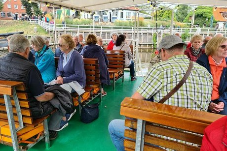 Menschen sitzen gemeinsam auf Holzbänken an Deck eines Ausflugsschiffs unter einer Markise.