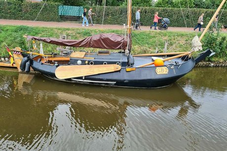 Ein schwarzes Plattbodenschiff mit hellen Seitenrudern aus Holz und dunkler Persenning liegt am Ufer des Kanals vertäut. Im Hintergrund die heckengesäumte Uferpromenade mit Spaziergängern.