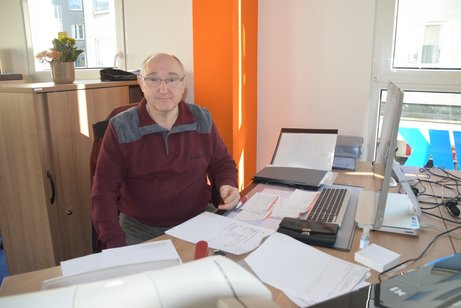 Roland Ruddat sitzt an einem Schreibtisch, auf welchem ein PC steht und viele Zettel liegen.