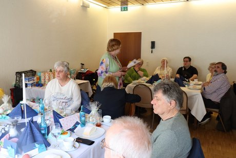 Eine Frau steht zwischen den Tischen, an denen mehrere Menschen sitzen und liest etwas vor.