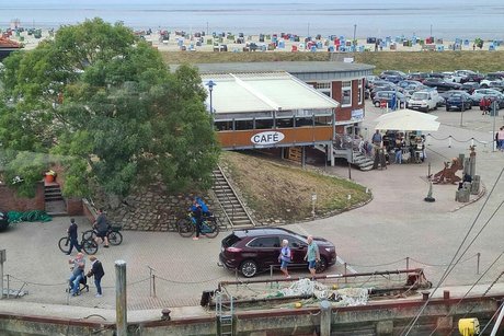 Blick von oben herab auf einen großen, mit Bäumen und einem Café bestandenen Platz. Im Hintergrund der Deich und dahinter ein mit bunten Strandkörben vollgestellter Strand und das Meer.