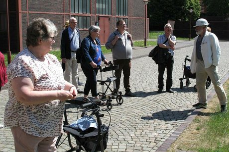 Eine Gruppe Menschen, einige davon mit Rollator stehen auf dem mit Kopfsteinen gepflasterten Zuweg zum Zechengelände