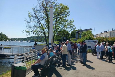 Eine Gruppe Menschen steht oder sitzt auf Bänken entlang des Ufers vom Baldeney-See in Essen. Im Hintergrund der weißgestrichene Bootsanleger der Weißen Flotte.