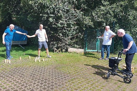 Zwei Männer stehen auf Rasenpflastersteinen und spielen ein finnisches Holzwurfspiel (Mjöllky). Rechts im Bild schauen ein Mann an einem Stehtisch und ein Mann mit Rollator den Spielenden zu.