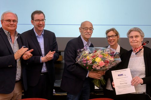 Drei Männer und zwei Frauen stehen auf einer Bühne. Eine Frau übergibt einem Mann einen Blumenstrauß, die andere Frau zeigt eine Urkunde mit der Aufschrift "Ehrenvorsitzender" in die Kamera.