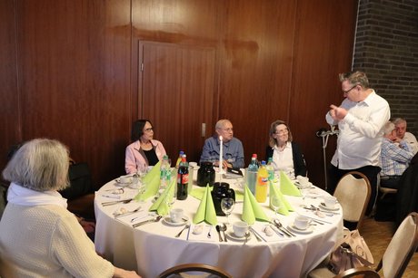 Mehrere Personen sitzen an einem runden Tisch und ein Mann steht daneben und unterh&auml;lt sich mit den Teilnehmern.