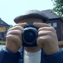 Die Skulptur Alltagsmensch in Straelen zeigt eine Person mit grauer Kappe, die einen Fotoapparat vor das Gesicht hält.