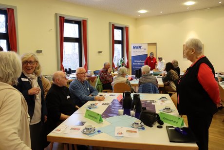 Mehrere Personen sitzen an Tischen und dazwischen stehen andere Personen und unterhalten sich.