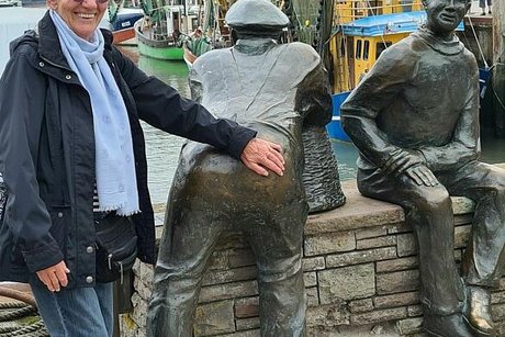 Zwei Fischer aus Bronze sind als Skulptur an der Hafenmauer arrangiert. Einer sitzt und einer lehnt sich über die Mauer und schaut auf die Fischkutter, die unterhalb im Wasser liegen. Eine Frau links im Bild fasst der Fischerskulptur scherzhaft an den Po.