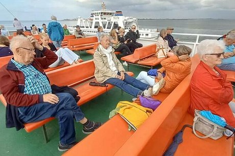 Menschen sitzen auf orangen Plastikbänken auf dem Außendeck einer Fähre.