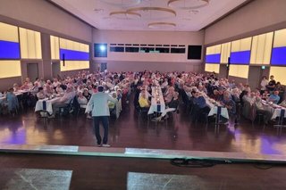 Viele Menschen sitzen in einem Saal an festlich gedeckten, langen Tischen.