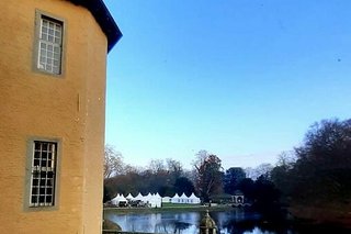 Blick von der Schlossterrasse über die in der Dämmerung schwarzglänzende, spiegelglatte Oberfläche des Burggrabens bis zur Parkwiese, auf der zahlreiche weiße Pavillons aufgebaut sind.