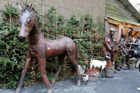 Ein fast lebensgroßes Pferd aus zusammengeschweißten Metallstücken steht vor einer Wand aus Tannenbäumen. Dahinter erkennt man weitere Metallskulpturen.