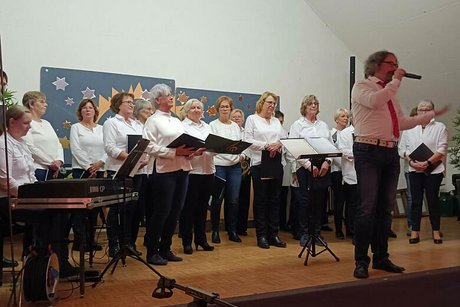Der Chorleiter spricht in ein Mikrofon. Hinter ihm steht eine große Gruppe an Frauen mit Notenheften in der Hand.