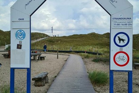 Ein großer Bogen mit der Aufschrift "Willkommen am Oase-Strand" spannt sich über einen Weg aus Holzbohlen, der durch Dünen zum Strand führt.